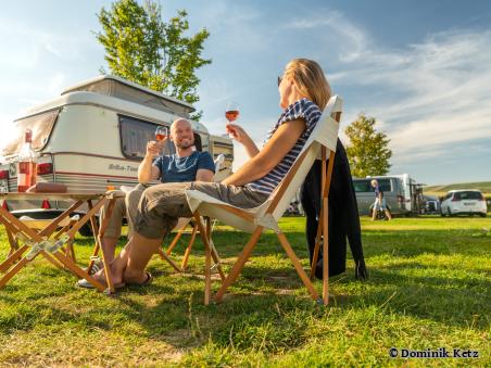 Ein Paar am Campingtisch mit einem Glas Wein vor ihrem Wohnwagen auf einer Campingwiese - Foto von Dominik Ketz