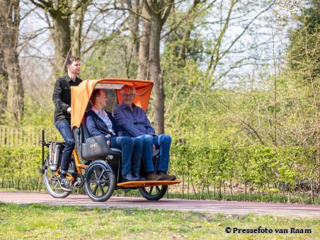 Ein Paar wird auf einer E-Rikscha von einem jungen Mann durch einen Park mit Bäumen gefahren - Foto von Pressefoto van Raam